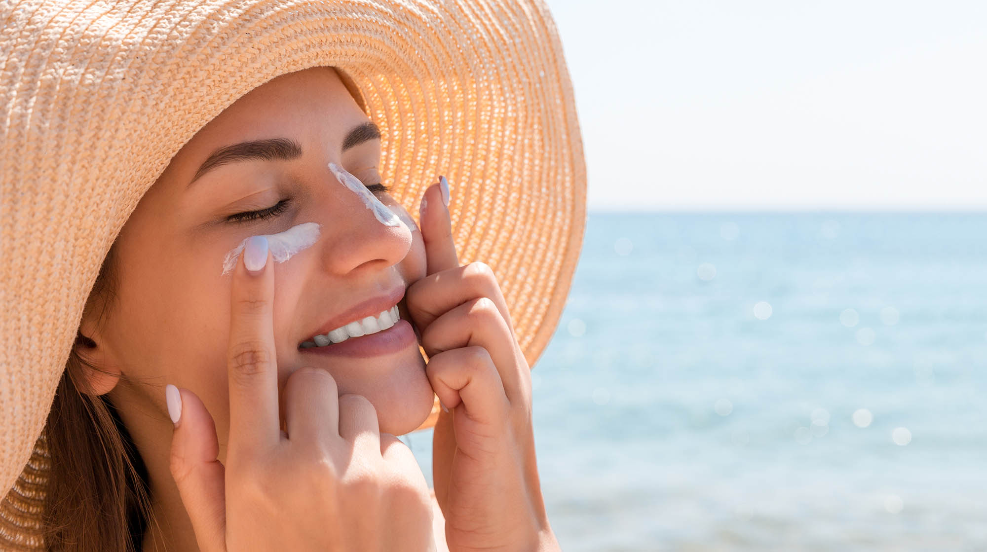 Schöne Frau am Strand mit Sonnenhut verteilt Sonnencreme im Gesicht